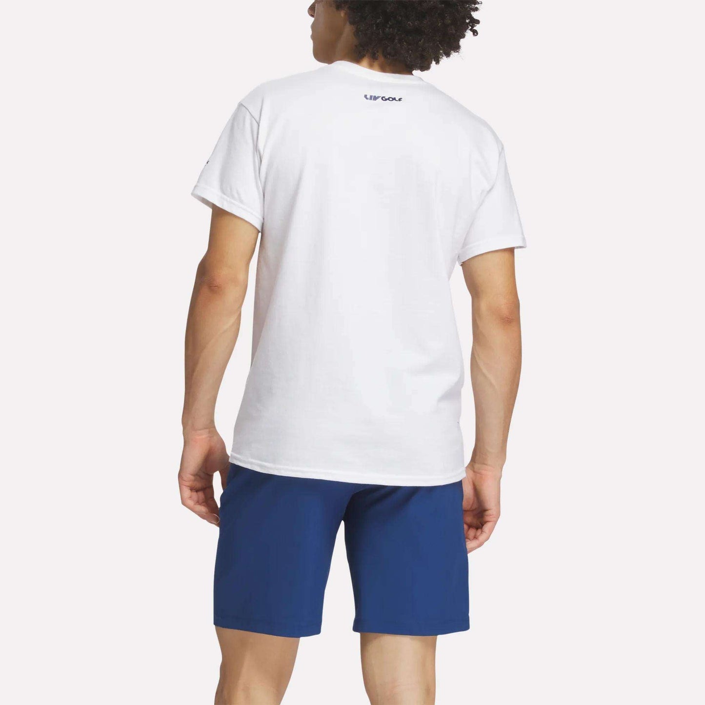 A person with curly hair is shown from the back wearing a plain white Reebok Men's Crushers Wordmark Tee and blue shorts. The background is bright and uncluttered. The word REEBOK is printed on the upper back of the T-shirt.