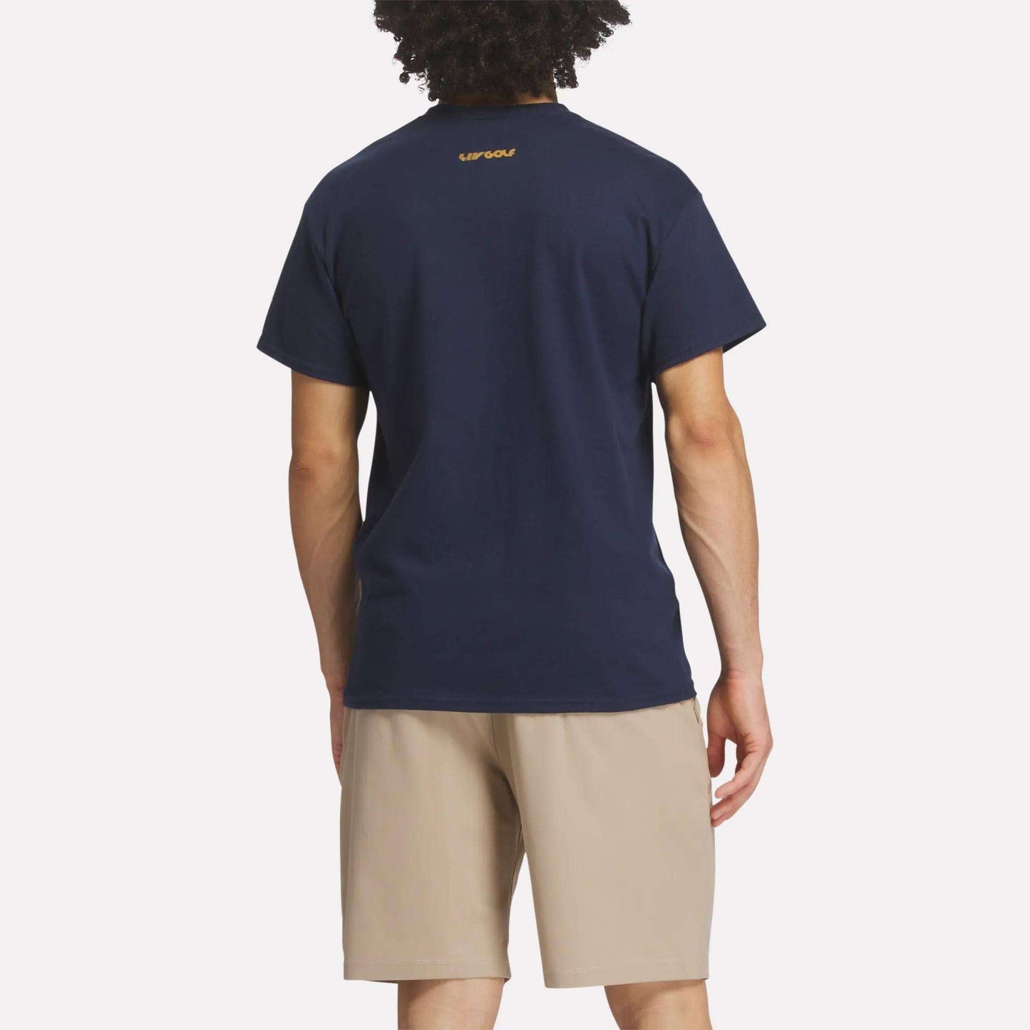 A person with curly hair is seen from behind, wearing the Reebok Men's Crushers Stacked Logo Tee in navy blue with a small yellow logo near the collar and light beige shorts, standing against a plain white background.