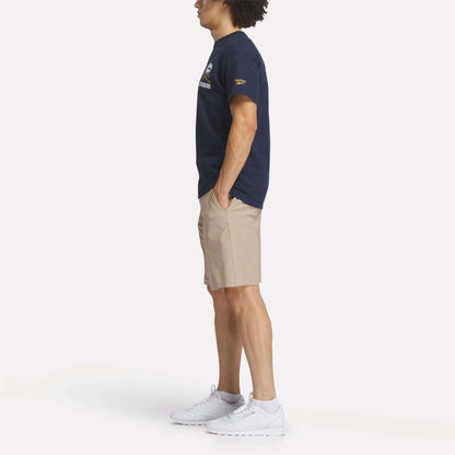 A person stands in profile wearing a navy Reebok Men's Crushers Stacked Logo Tee, beige shorts, and white sneakers, hands in pockets, against a plain white background.