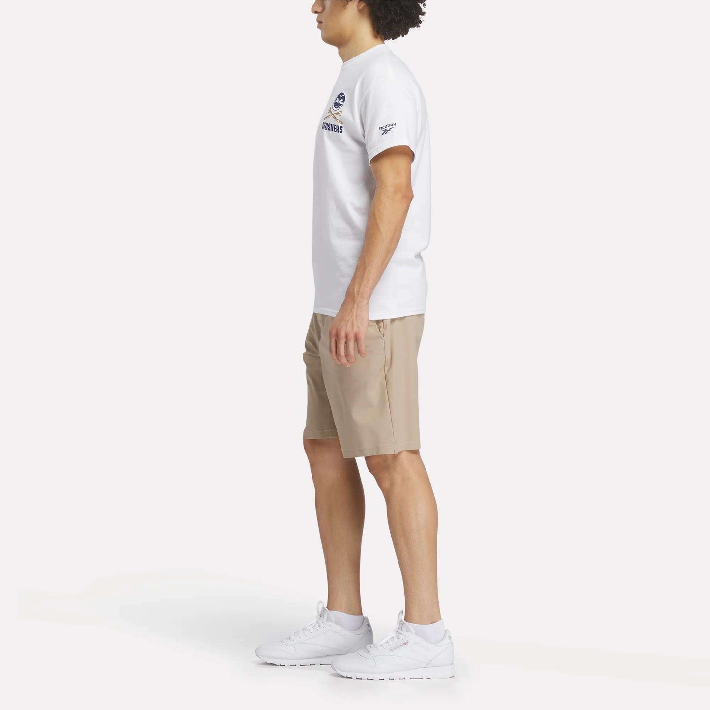 A man stands in profile against a plain white background wearing a Reebok Men's Crushers Stacked Logo Tee, beige shorts, and white Reebok sneakers.