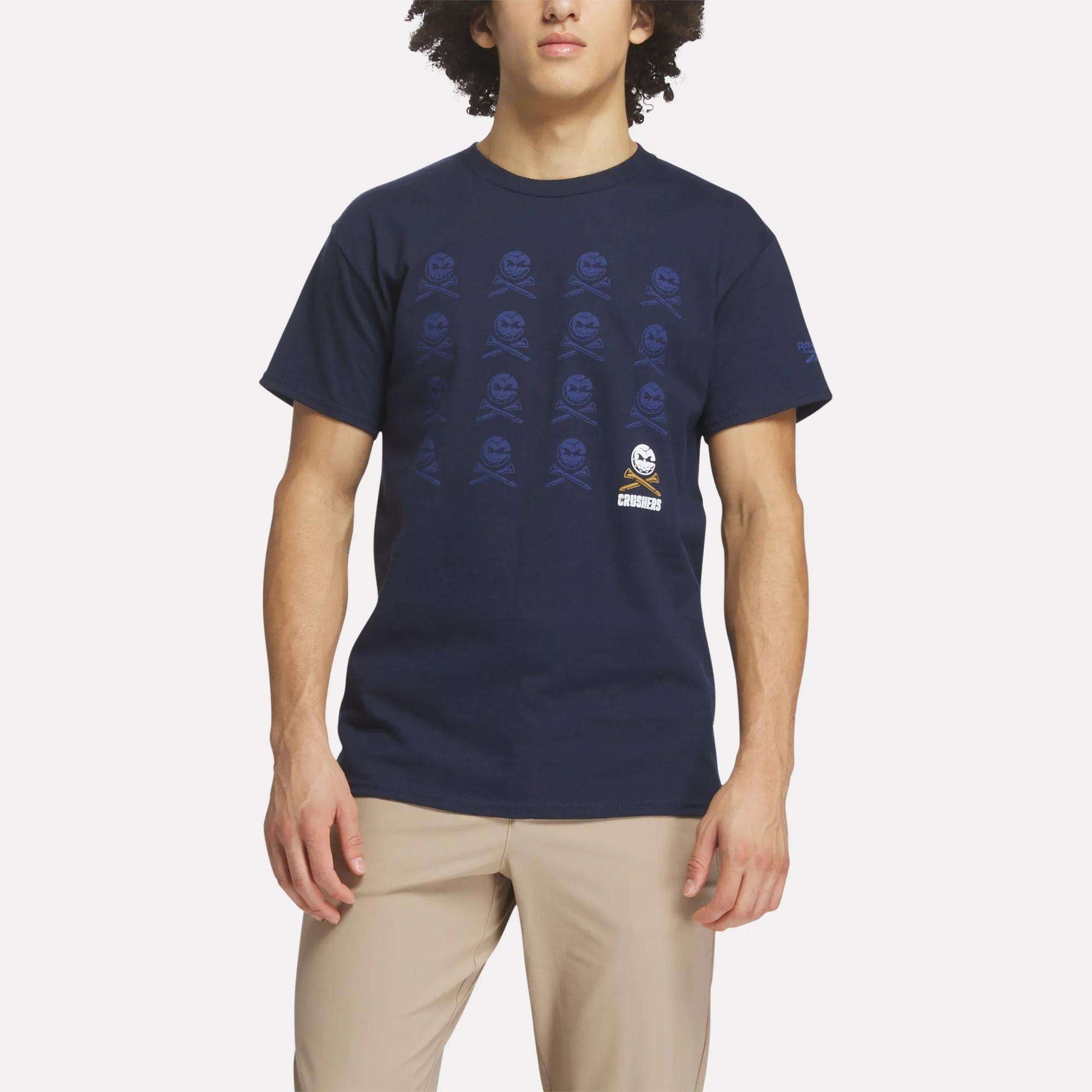 A person wearing the Reebok Men's Crushers Icon Tee in navy blue with a repeating skull and crossbones pattern and tan pants, standing against a plain light background.