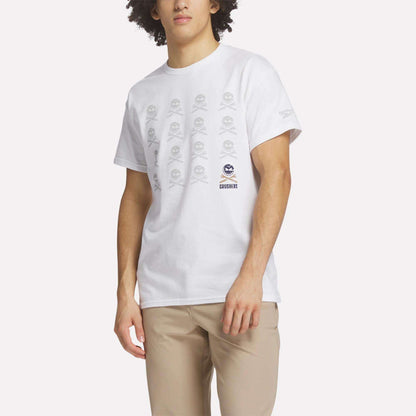A man wears the Reebok Men's Crushers Icon Tee, featuring repeated gray skull and crossbones graphics on white, along with beige pants, standing against a plain light background.