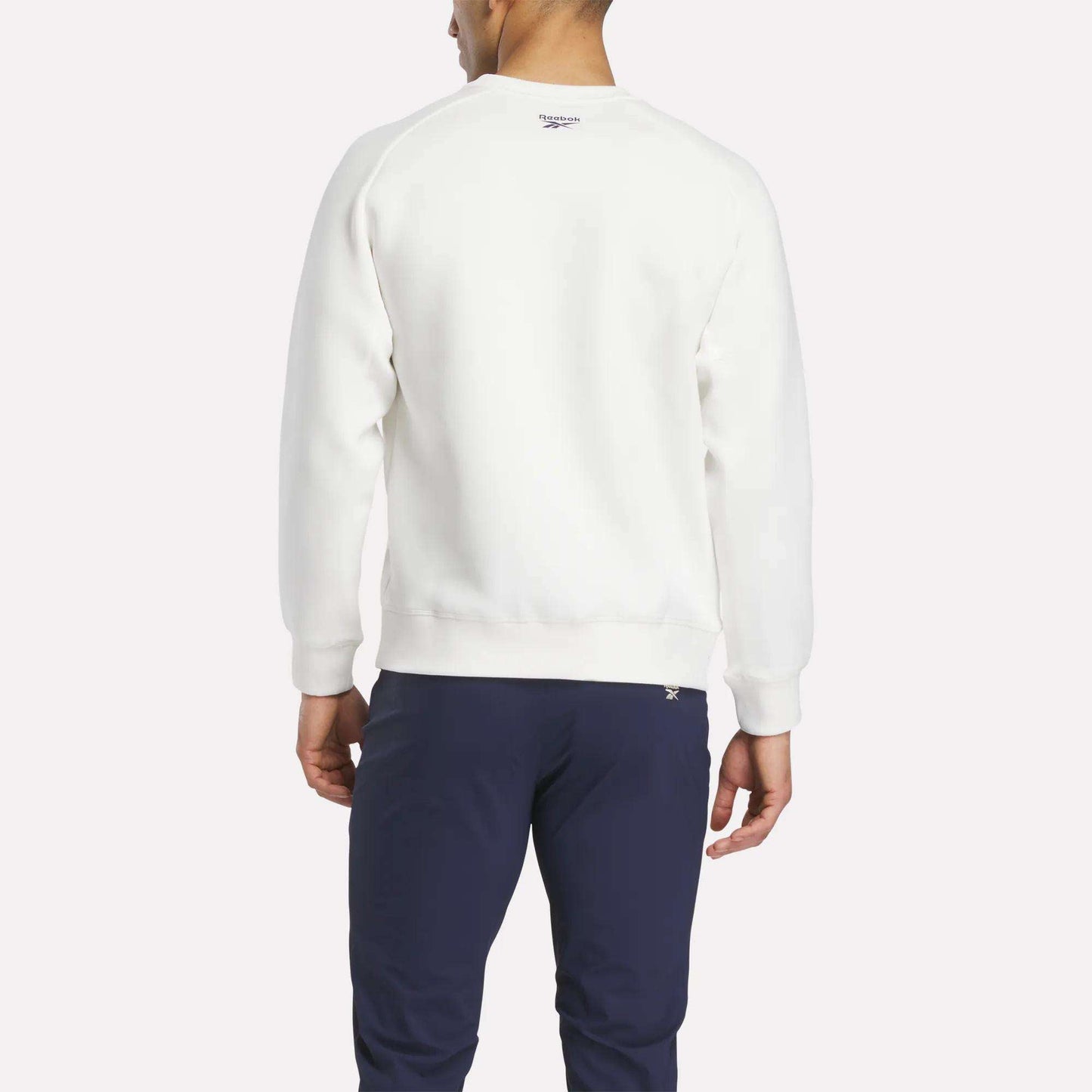 A man faces away from the camera, wearing a white Reebok Men's Scuba Knit Crew Sweatshirt and navy blue pants against a plain light background.