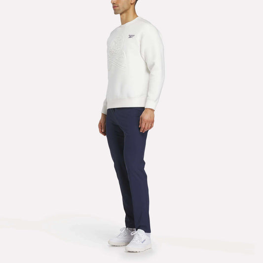 A man stands slightly to the side, wearing a white Reebok Men's Scuba Knit Crew Sweatshirt, navy blue pants, and white sneakers against a plain white background.