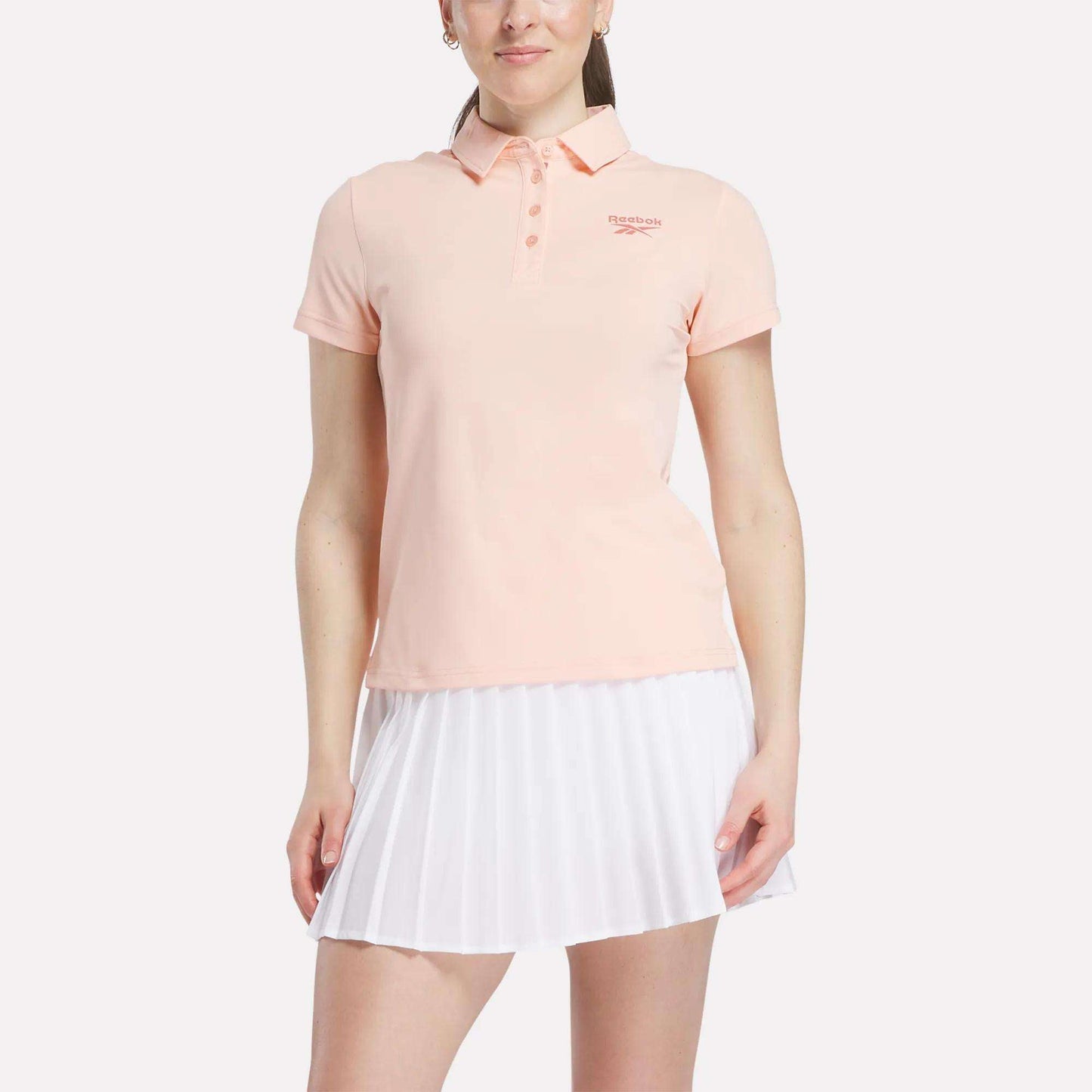 A woman stands against a white background wearing a light pink Reebok Women's Micro Pique Polo Shirt with a small left chest logo and a white pleated skirt, both made from stretch fabric for comfort.