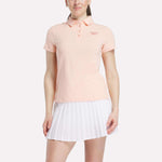 A woman stands against a white background wearing a light pink Reebok Women's Micro Pique Polo Shirt with a small left chest logo and a white pleated skirt, both made from stretch fabric for comfort.