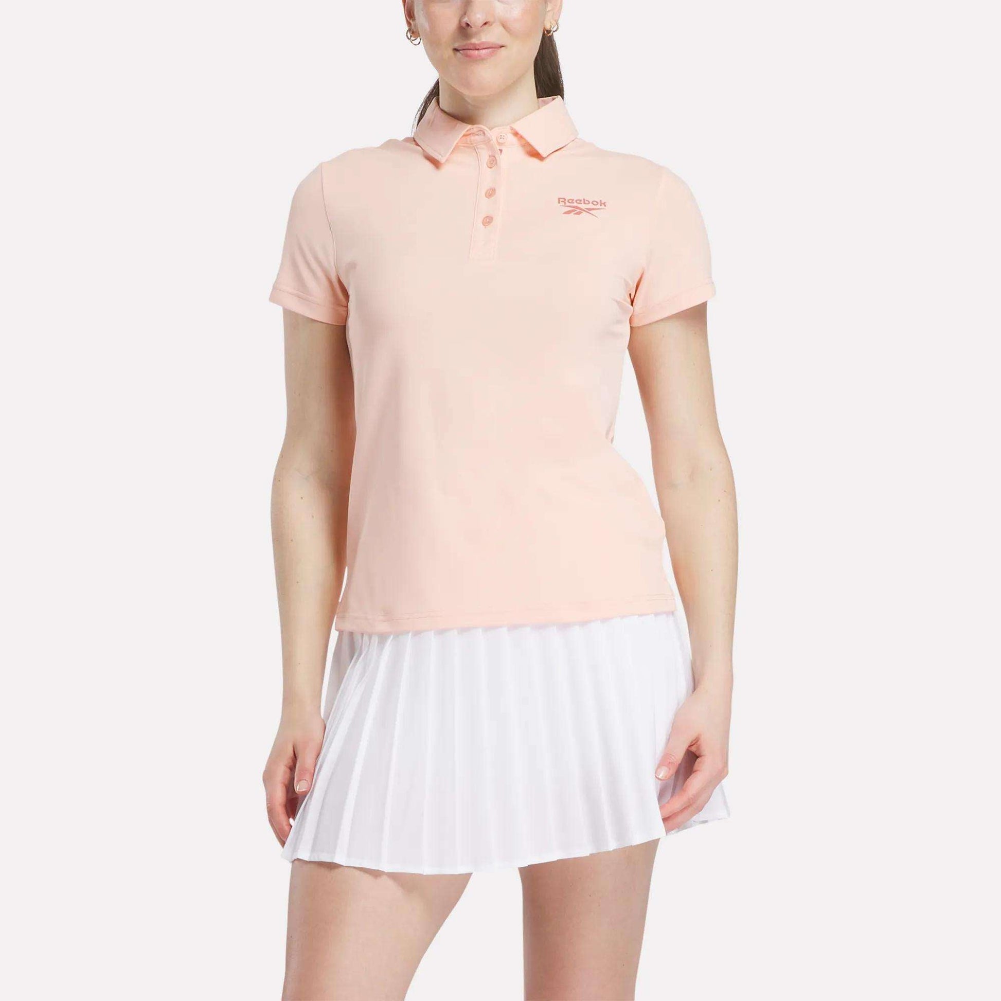 A woman stands against a white background wearing a light pink Reebok Women's Micro Pique Polo Shirt with a small left chest logo and a white pleated skirt, both made from stretch fabric for comfort.