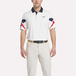 A man in a Reebok Men's Vector Sleeve Polo Shirt with moisture-wicking fabric, navy and red sleeve and collar accents, light-colored pants, and a black belt stands against a plain background.