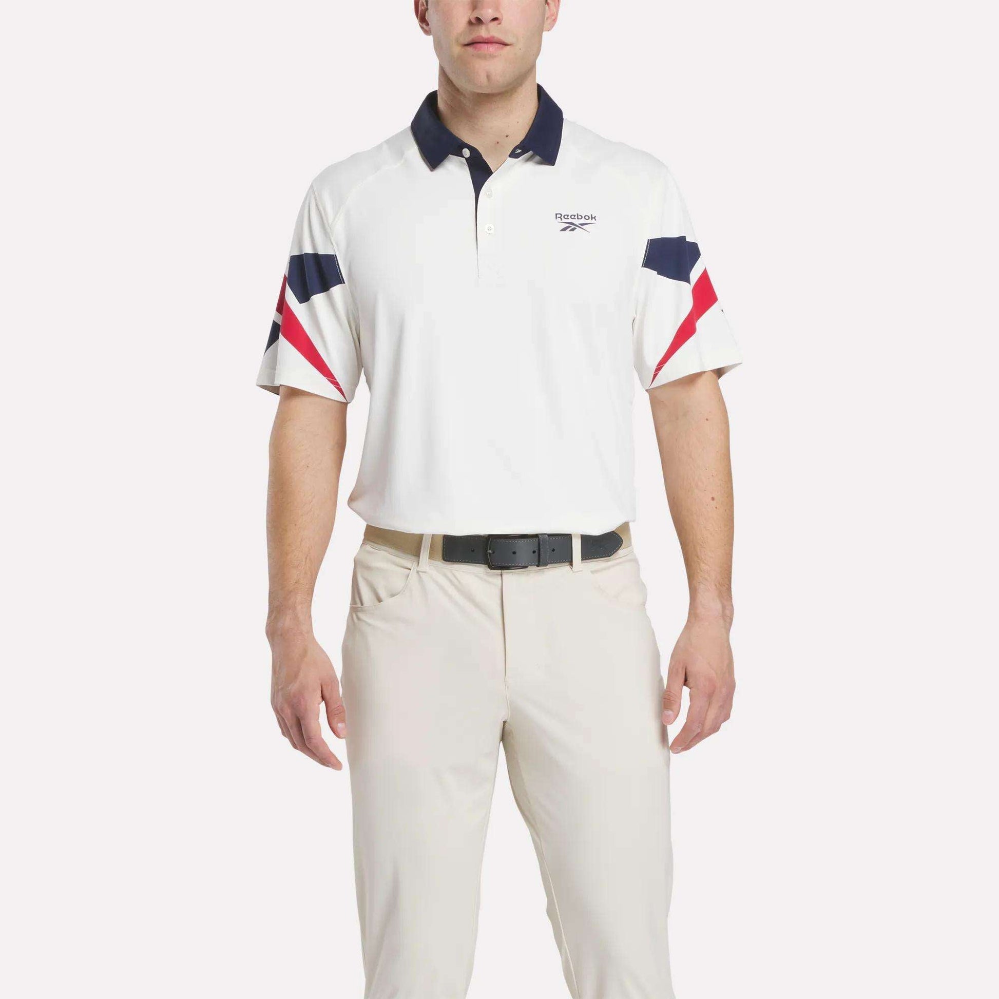 A man in a Reebok Men's Vector Sleeve Polo Shirt with moisture-wicking fabric, navy and red sleeve and collar accents, light-colored pants, and a black belt stands against a plain background.