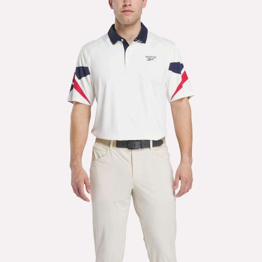 A man in a Reebok Men's Vector Sleeve Polo Shirt with moisture-wicking fabric, navy and red sleeve and collar accents, light-colored pants, and a black belt stands against a plain background.