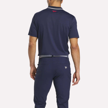 A man stands with his back to the camera, wearing a Reebok Men's Vector Argyle Print Polo in navy and matching navy pants with a light gray belt, both crafted from stretch knit fabric for superior comfort.