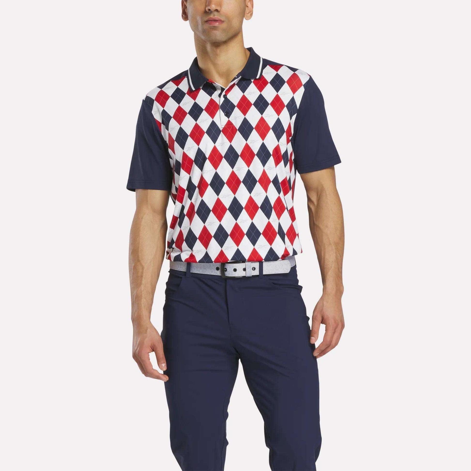 A man wears the Reebok Men's Vector Argyle Print Polo, featuring a red, white, and blue argyle pattern on stretch RBK-DRY fabric, with navy pants and a light gray belt, standing against a plain white background.