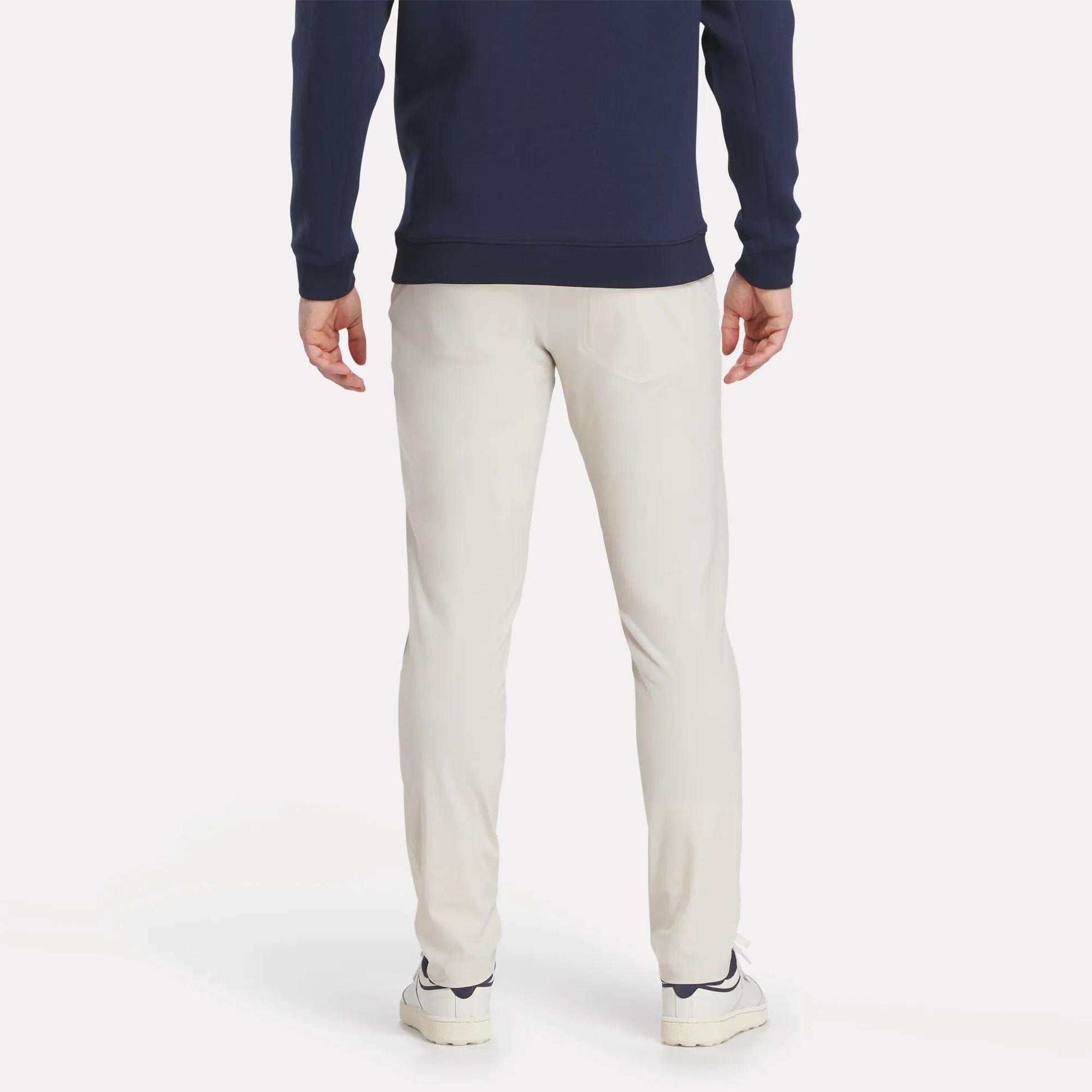 A person stands with their back to the camera, dressed in Reebok's Men's Favorite 5-Pocket Relaxed Fit Golf Pants with RBK-DRY tech, a navy long-sleeve top, and white sneakers against a plain white background.