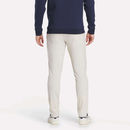 A person stands with their back to the camera, dressed in Reebok's Men's Favorite 5-Pocket Relaxed Fit Golf Pants with RBK-DRY tech, a navy long-sleeve top, and white sneakers against a plain white background.