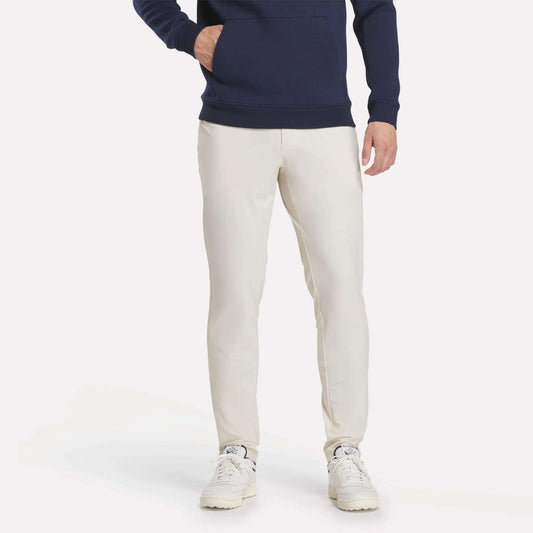 A person wearing a navy hoodie, Reebok Men's Favorite 5-Pocket Relaxed Fit Golf Pants in light beige, and white sneakers stands against a plain white background with one hand in their pocket. Their head is not visible in the image.