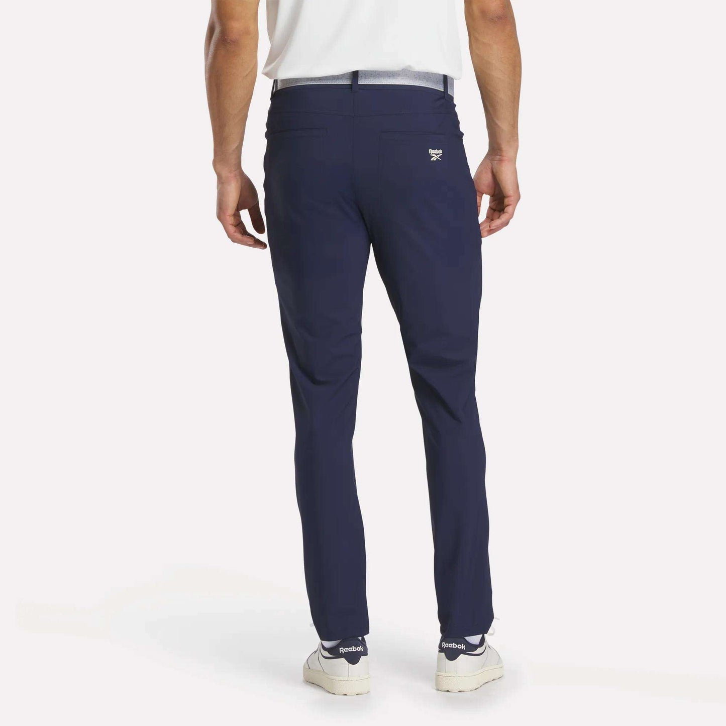 A man faces away from the camera, wearing Reebok Men's Favorite 5-Pocket Relaxed Fit Golf Pants in wrinkle-resistant navy blue, paired with a white shirt and sneakers against a plain white background.