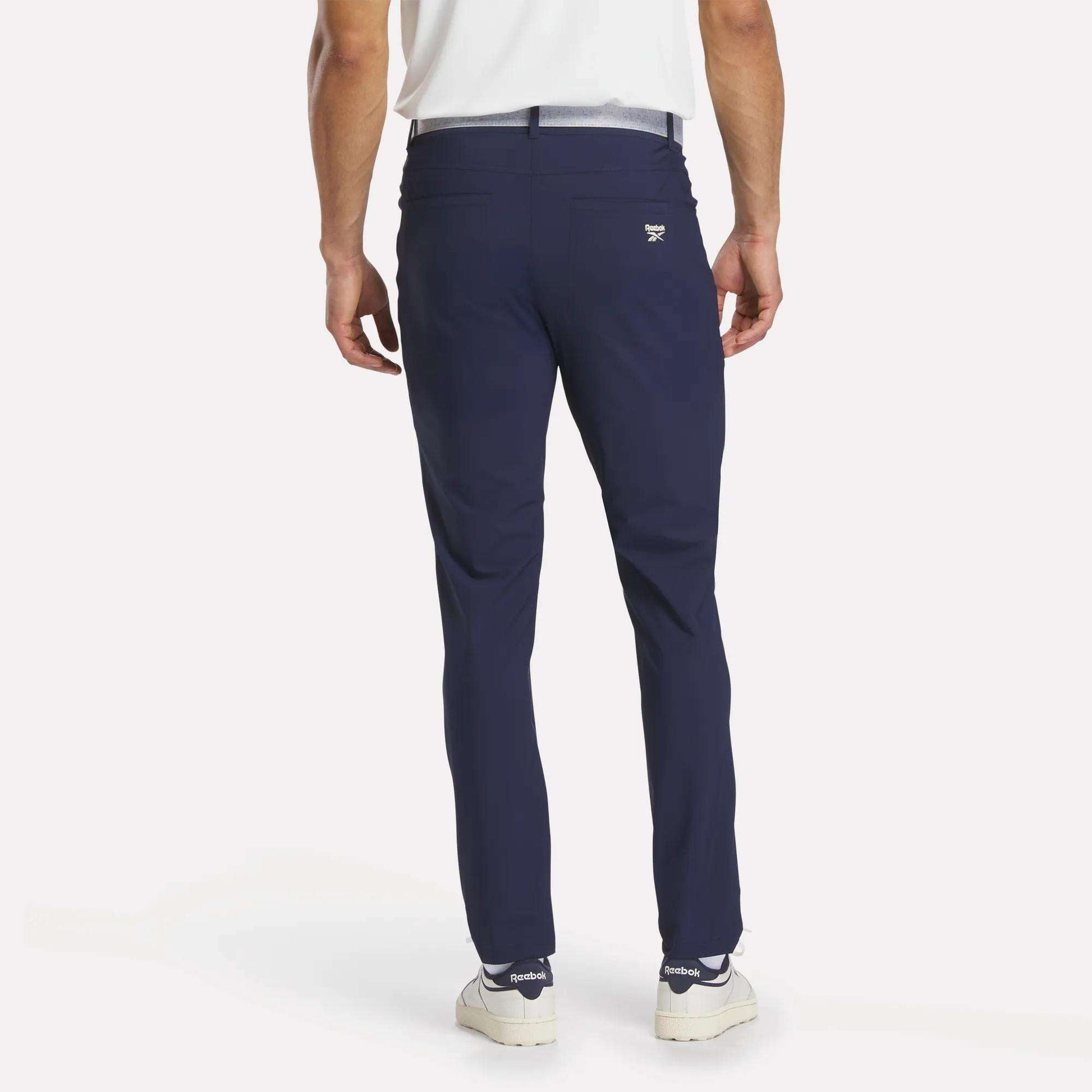A man faces away from the camera, wearing Reebok Men's Favorite 5-Pocket Relaxed Fit Golf Pants in wrinkle-resistant navy blue, paired with a white shirt and sneakers against a plain white background.