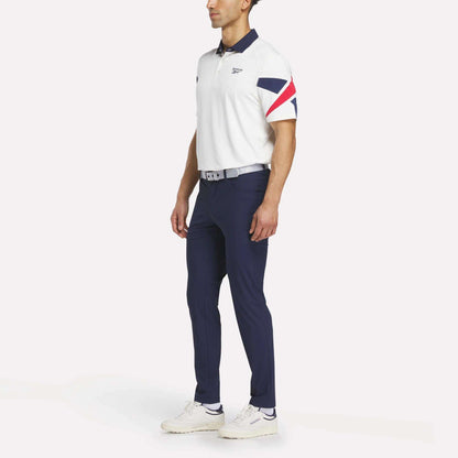 A man in a white polo with red and navy accents, Reebok Men's Favorite 5-Pocket Relaxed Fit Golf Pants in navy, a light grey belt, and white sneakers stands slightly turned against a plain background.