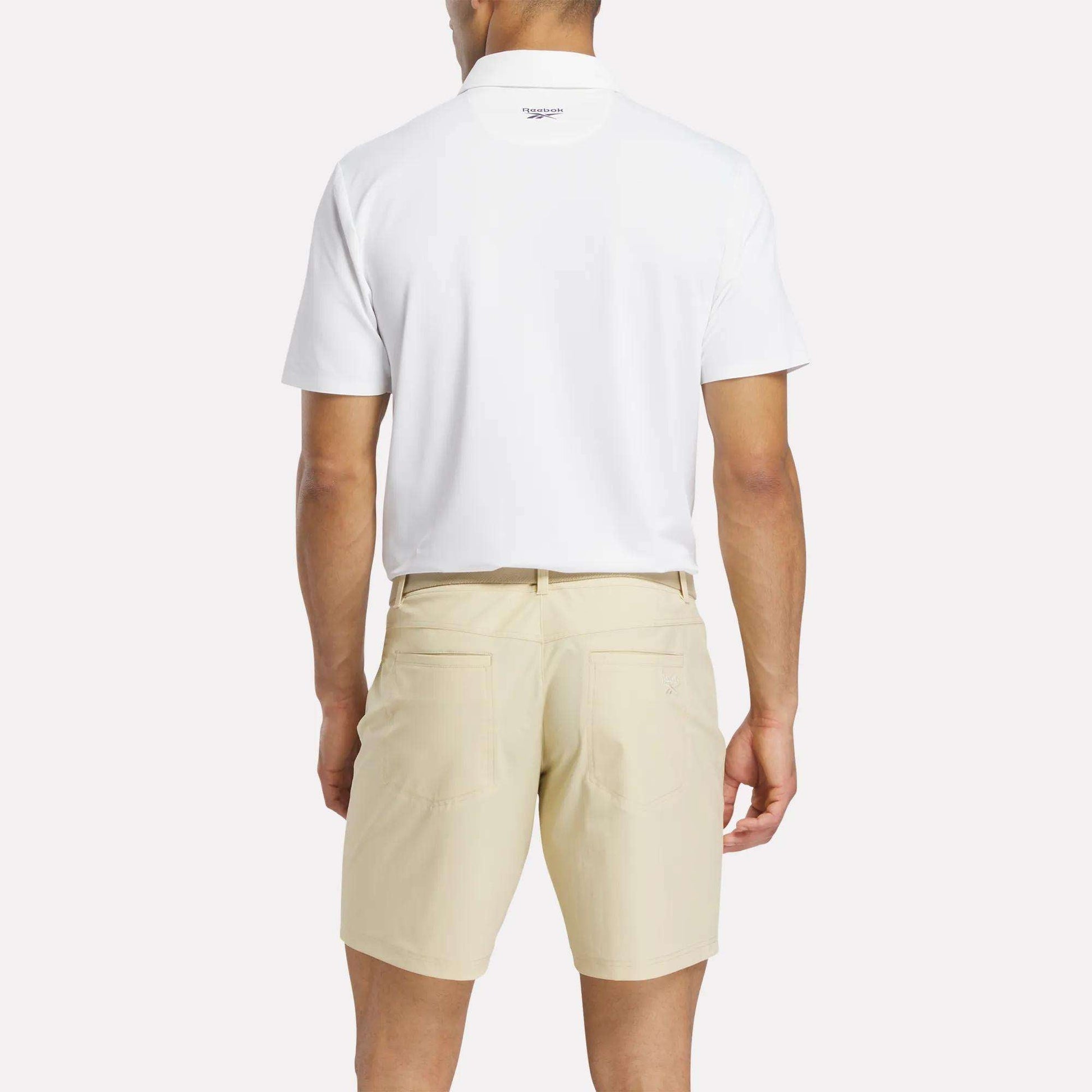 A man faces away from the camera, wearing a white Reebok Men's Bonded Pique Polo Shirt and light beige shorts against a plain background.
