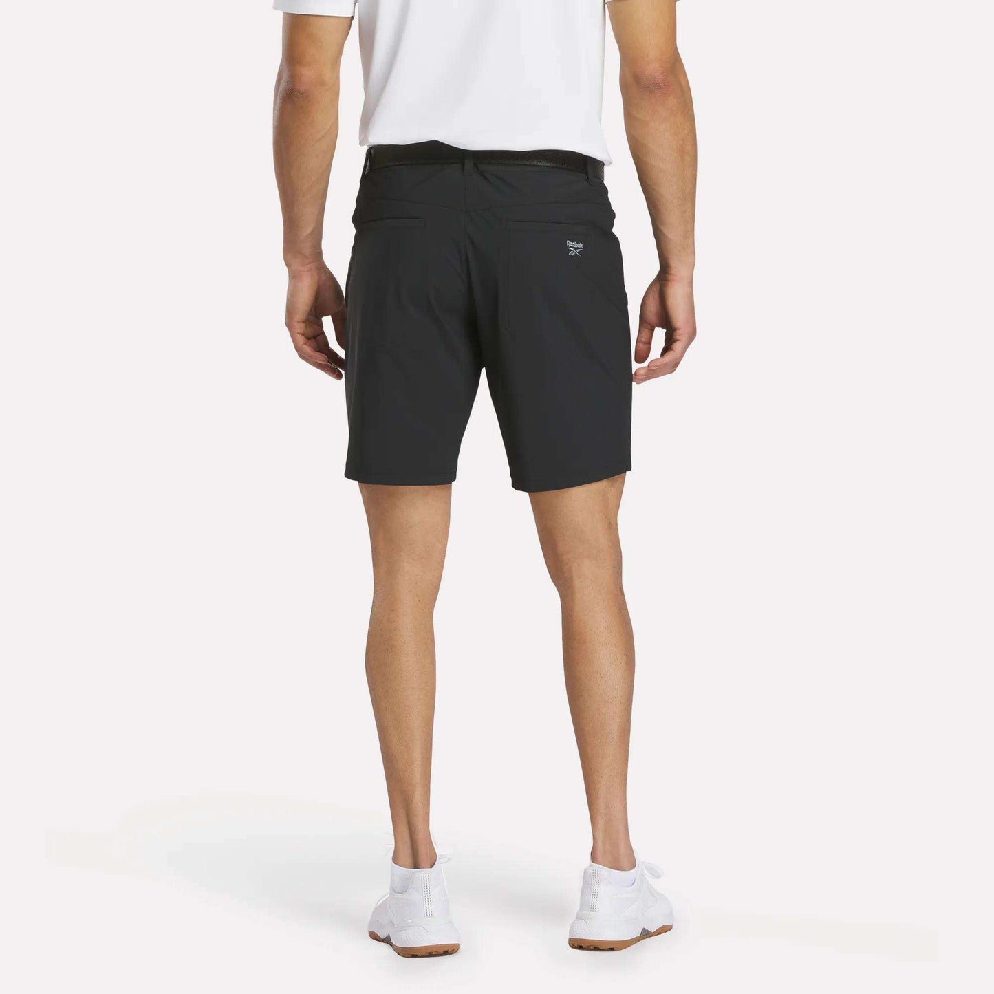A person stands facing away from the camera, wearing a white polo shirt, Reebok Men's Favorite Golf Shorts 8.5", and white sneakers against a plain background.