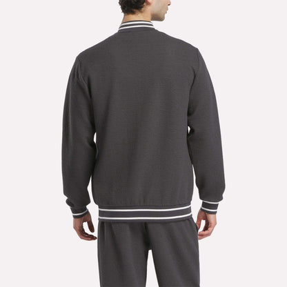 A man is seen from behind wearing the Reebok Men's Club Sport Textured Jacket in dark gray, featuring white and gray striped cuffs, collar, and waistband. Matching pants complete the sporty look against a plain white background.