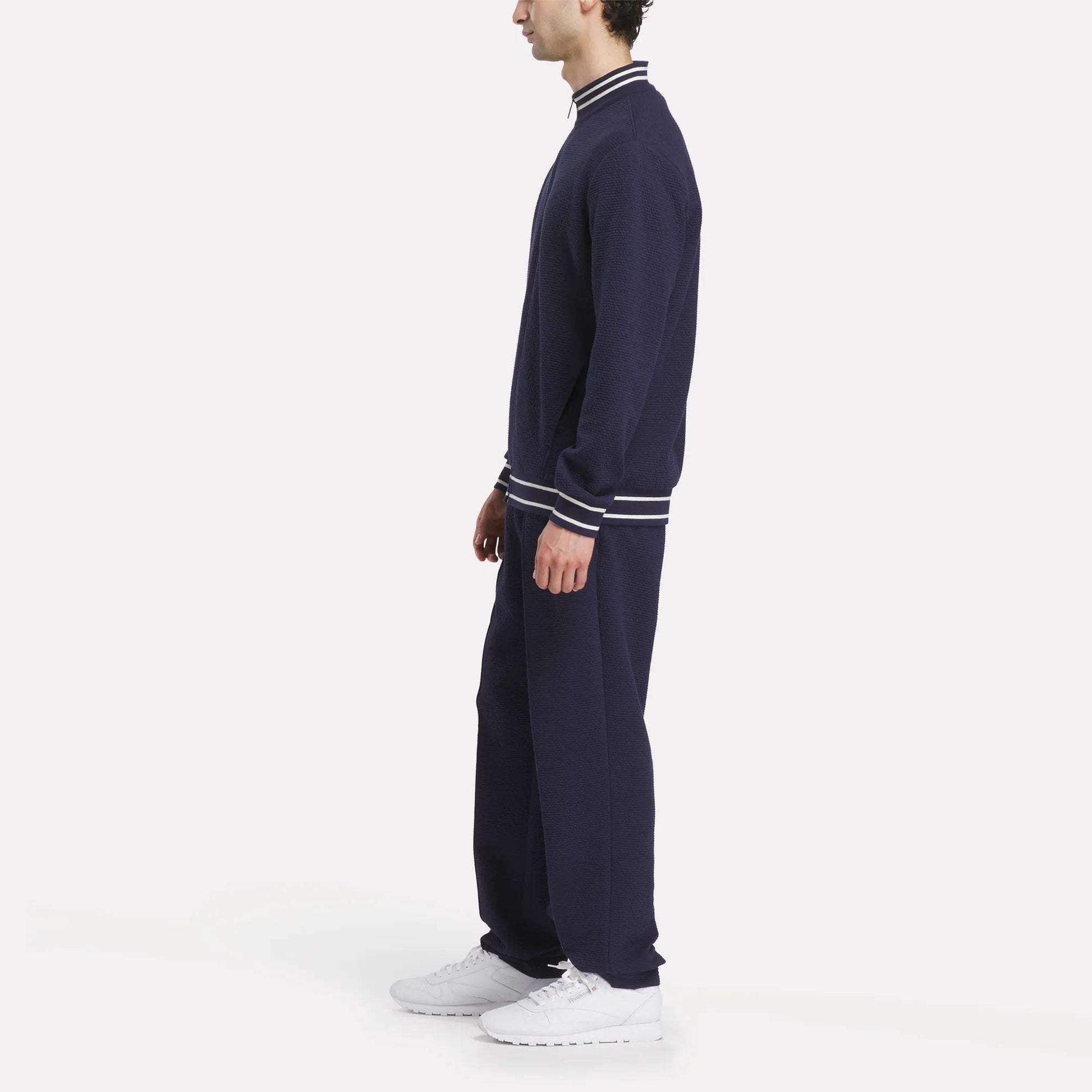 A man in profile wears a sporty navy tracksuit with white-striped cuffs and collar, featuring the Reebok Men's Club Sport Textured Jacket and white sneakers, standing against a plain white background.