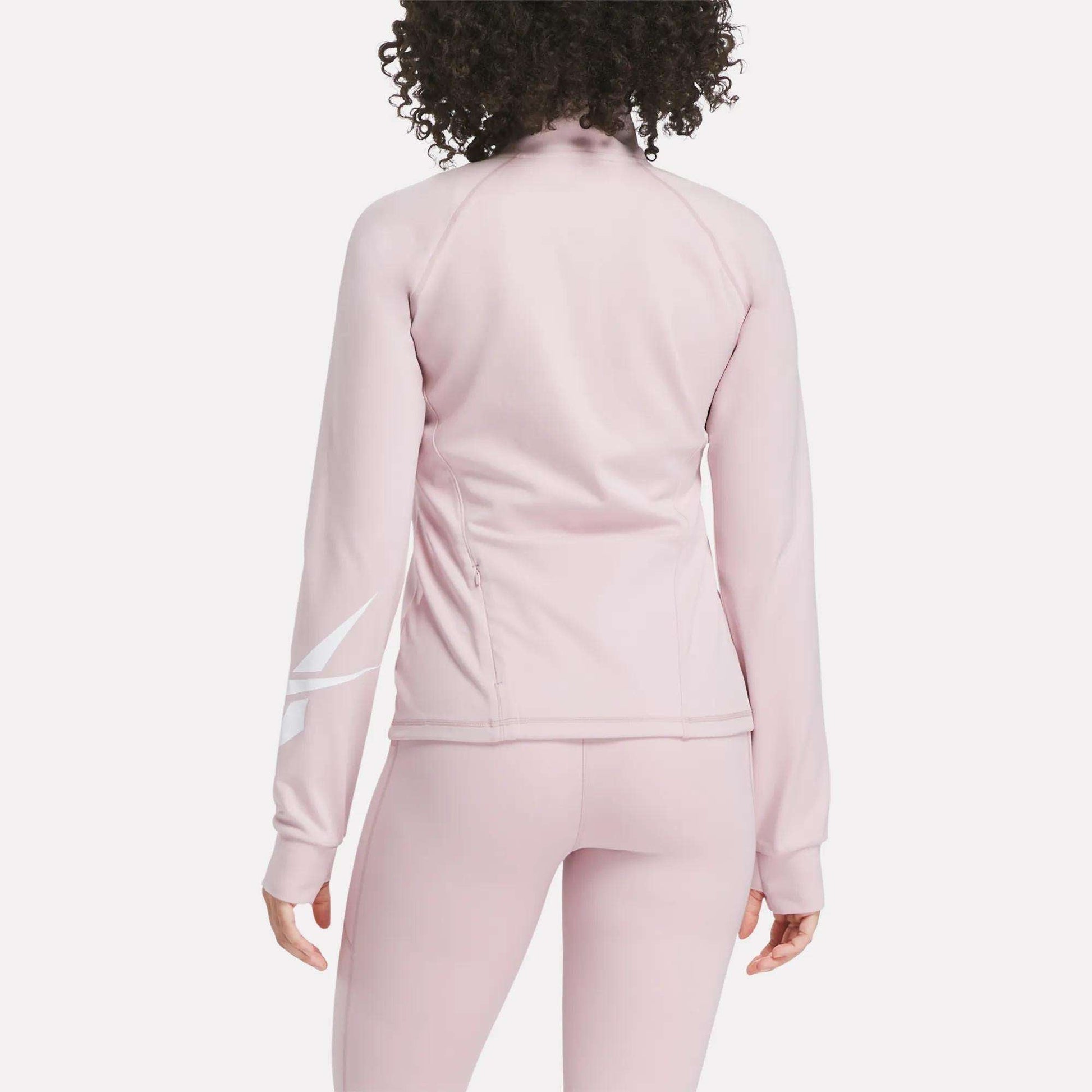 A woman with curly hair is seen from the back wearing the Reebok Women's Motion Core Full-Zip Hoodie in light pink with a white diagonal design on the right sleeve—perfect for workouts. Matching leggings complete her athletic look.