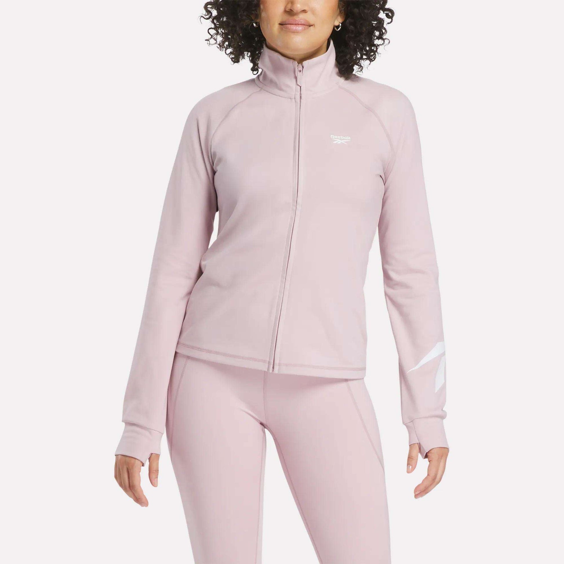A person wearing a light pink Reebok Women's Motion Core Full-Zip Hoodie and matching leggings, standing with arms relaxed by their sides against a plain white background.