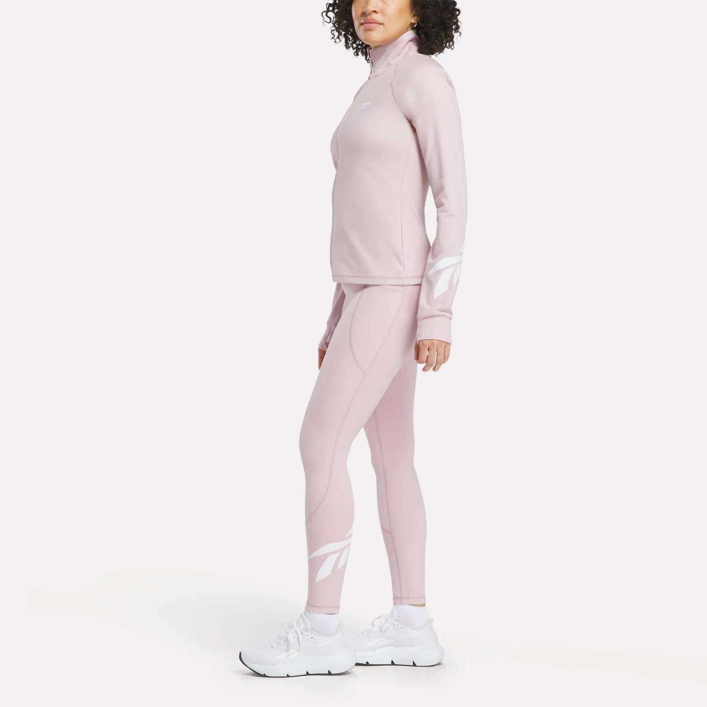 A woman stands in side profile against a white background wearing the Reebok Women's Motion Core Full-Zip Hoodie in light pink, styled with matching athletic pants and white sneakers. The hoodie features bold white logo designs on the sleeves and lower legs.