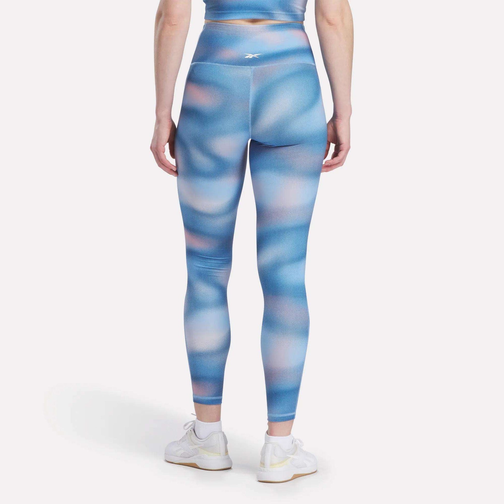 A woman faces away, wearing Reebok Women's Fluid Print High-Rise Tights with a matching top and white sneakers, all set against a plain white background.