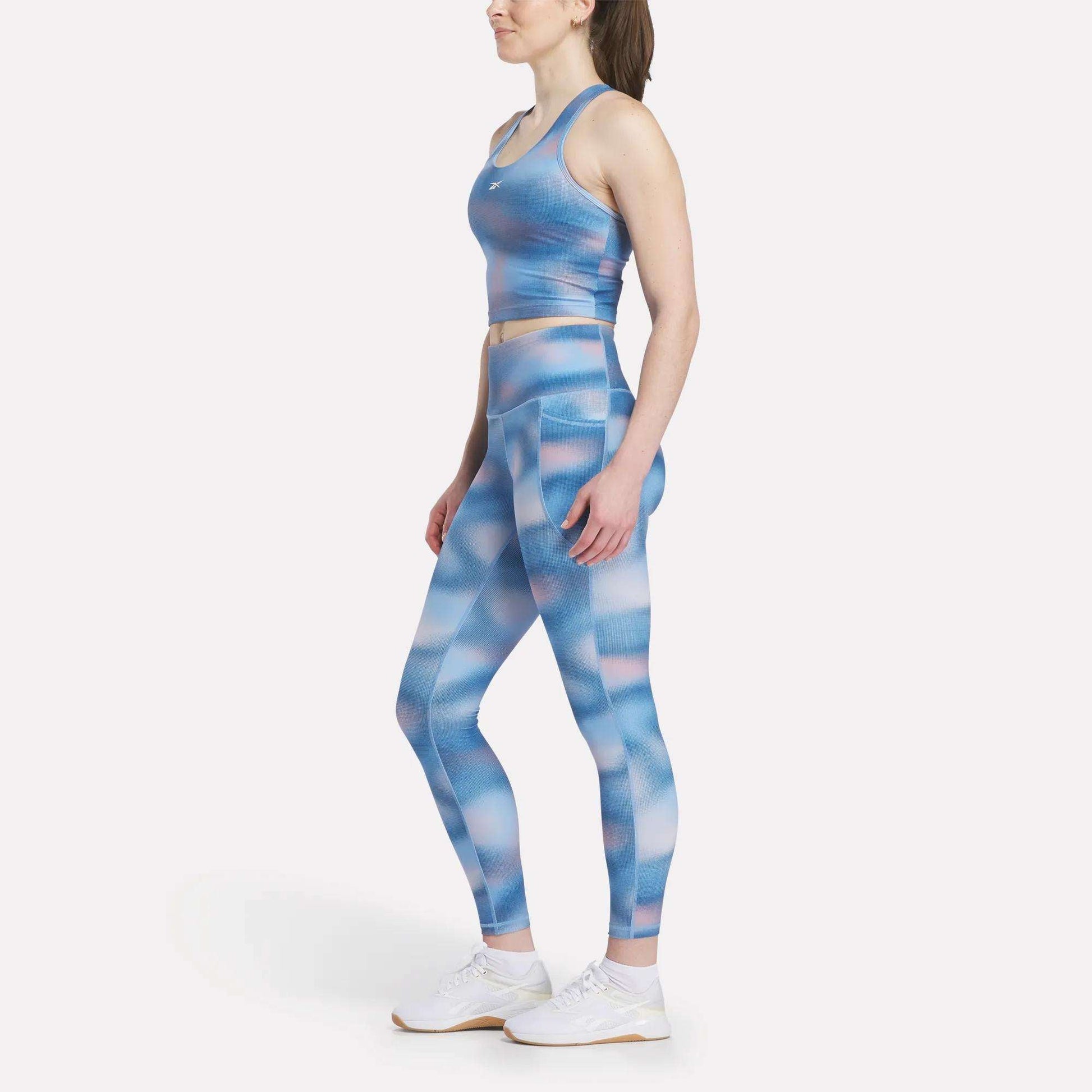 A woman stands in profile wearing Reebok Women's Fluid Print High-Rise Tights with a matching sports bra and white sneakers, highlighting the high-rise waist against a plain white background.