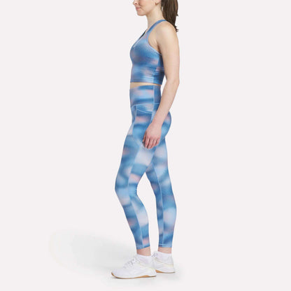 A woman in profile wears a Reebok Women's Cropped Bra Tank Top and matching blue-pink patterned leggings with light-impact support, paired with white athletic shoes, all set against a plain white background.