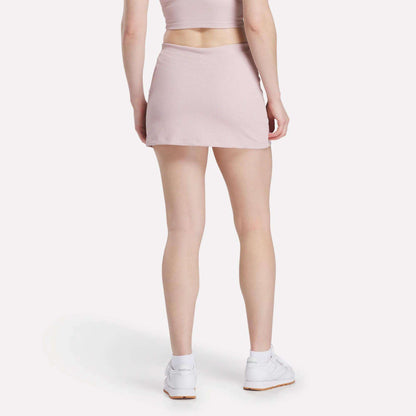 A woman stands with her back to the camera in a light pink Reebok Women's Time Out Slim Tennis Skort and matching crop top, styled with white sneakers, against a plain white background for a chic activewear look.