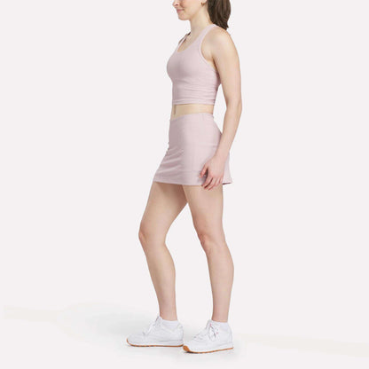 A woman in profile models a light pink sleeveless crop top with the women's Reebok Time Out Slim Tennis Skort and white Reebok sneakers, set against a plain white background.