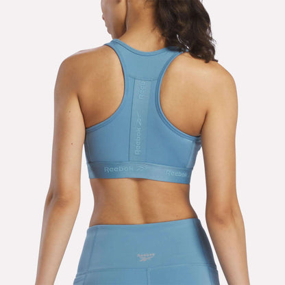 A woman is shown from the back wearing a light blue Reebok Women’s Medium-Impact Sports Bra with matching leggings. The sports bra features a racerback design, visible Reebok logo, and removable cups for customizable comfort.
