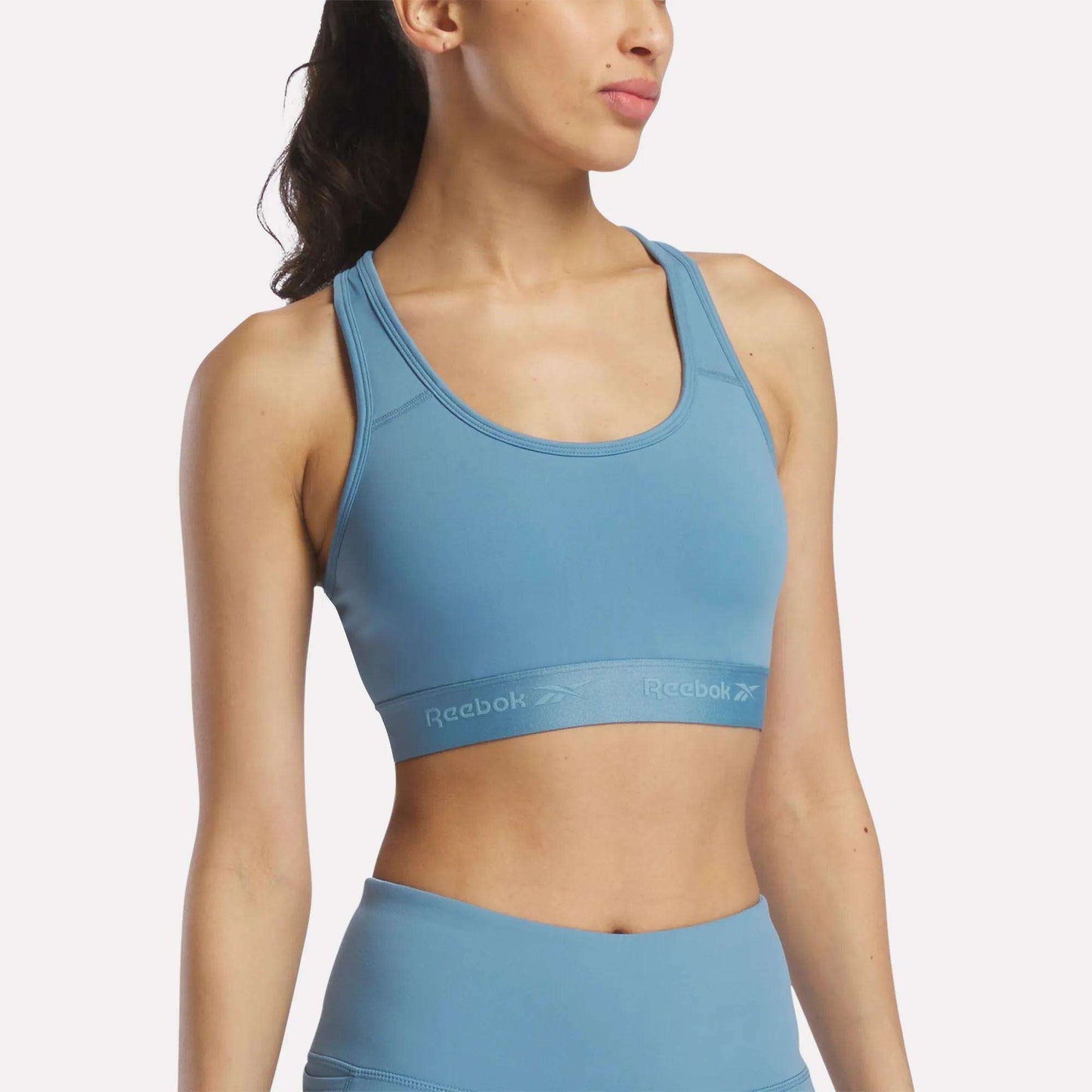 A woman stands against a plain background, facing slightly to the side with her head turned away from the camera, wearing a Reebok Women's Medium-Impact Sports Bra in Warped Blue with removable cups, paired with matching leggings.