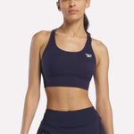 A woman stands against a plain light background wearing the Reebok Women's Rush Medium-Impact Sports Bra in navy blue with matching bottoms, ideal for medium-impact workouts.
