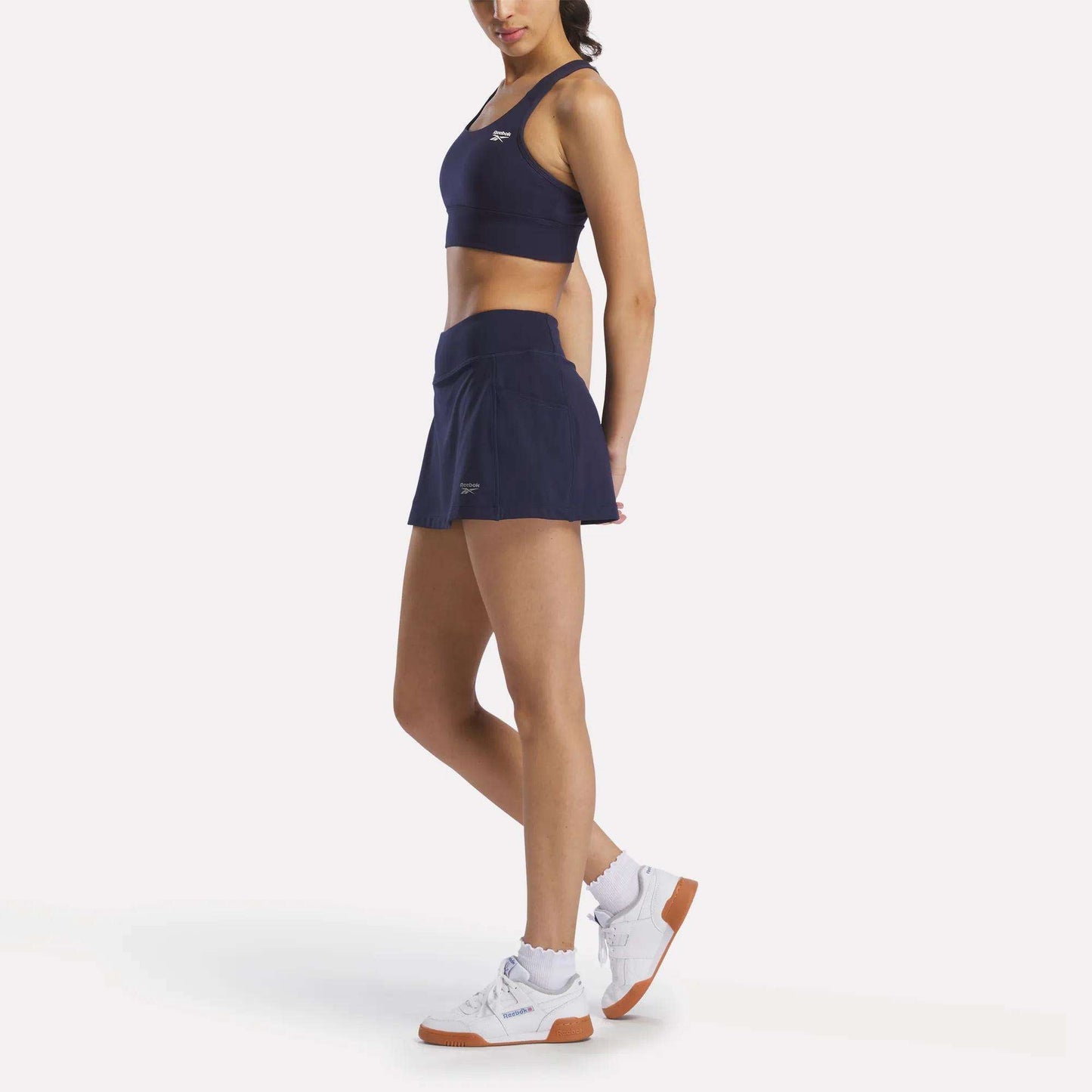 A woman wears a Reebok Women's Rush Medium-Impact Sports Bra in navy blue, a matching tennis skirt, white socks, and white athletic shoes while standing against a plain white background.