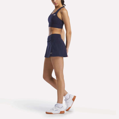 A woman wears a Reebok Women's Rush Medium-Impact Sports Bra in navy blue, a matching tennis skirt, white socks, and white athletic shoes while standing against a plain white background.