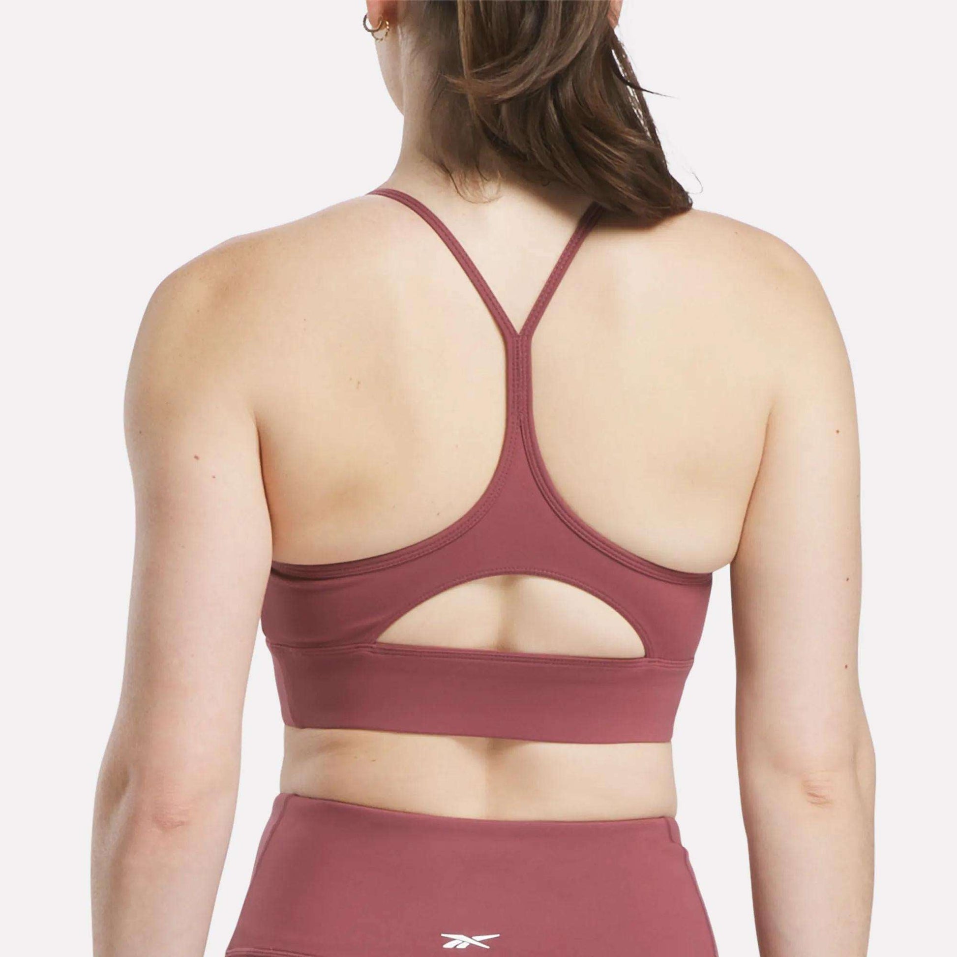 A woman is seen from behind wearing the Reebok Women's Back Bra in mauve with thin racerback straps and a small keyhole cutout, paired with matching high-waisted leggings.
