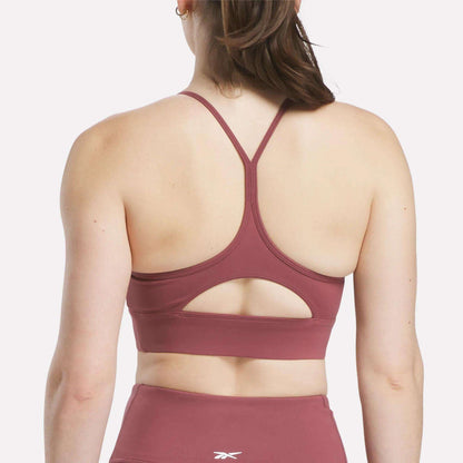 A woman is seen from behind wearing the Reebok Women's Back Bra in mauve with thin racerback straps and a small keyhole cutout, paired with matching high-waisted leggings.
