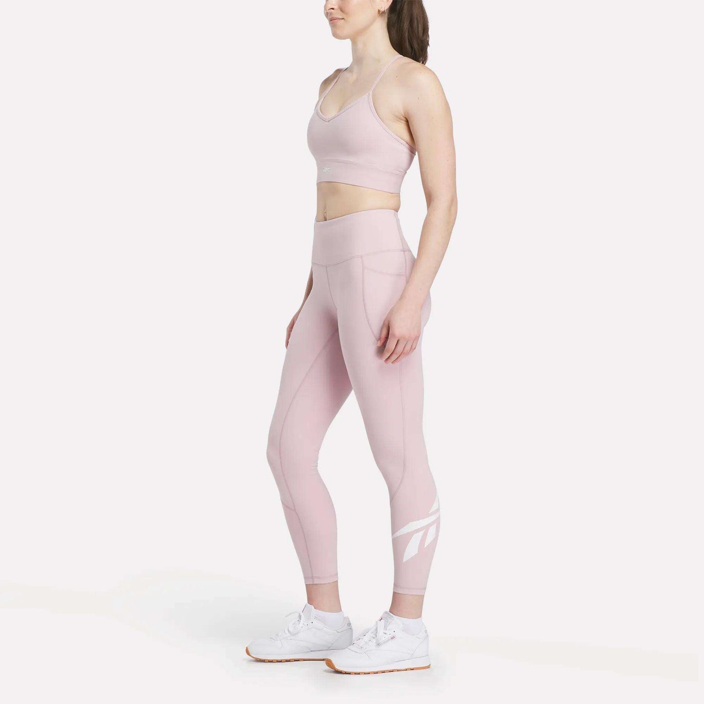 A woman models the Reebok Women's Back Bra and matching high-waisted leggings in light pink with white athletic shoes, standing sideways against a plain white background to highlight medium-impact support.