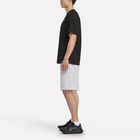 A man stands in profile facing left, wearing a Reebok Men's Director Short Sleeve Top, light gray athletic shorts, black sneakers, and white socks against a plain white background.