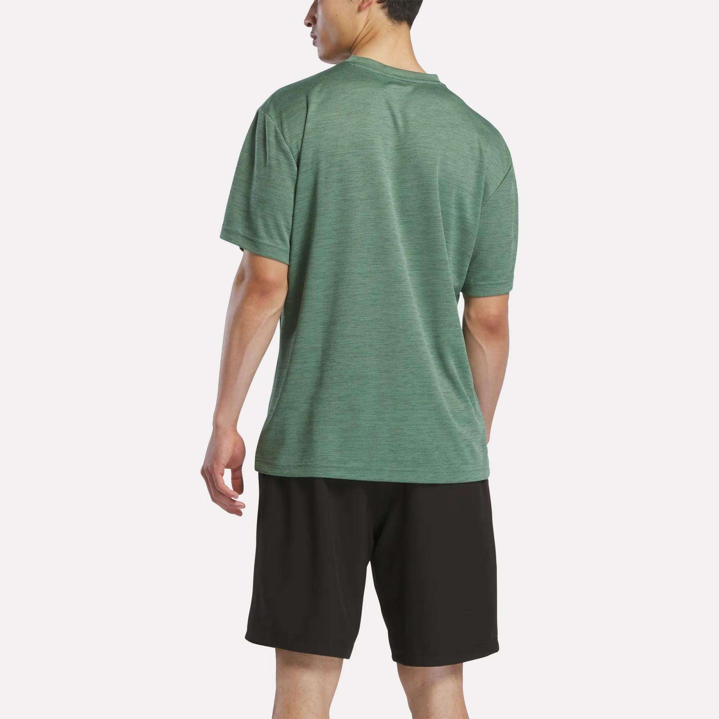 A man stands facing away, wearing a green Reebok Men's Director Short Sleeve Top and black shorts against a plain white background.