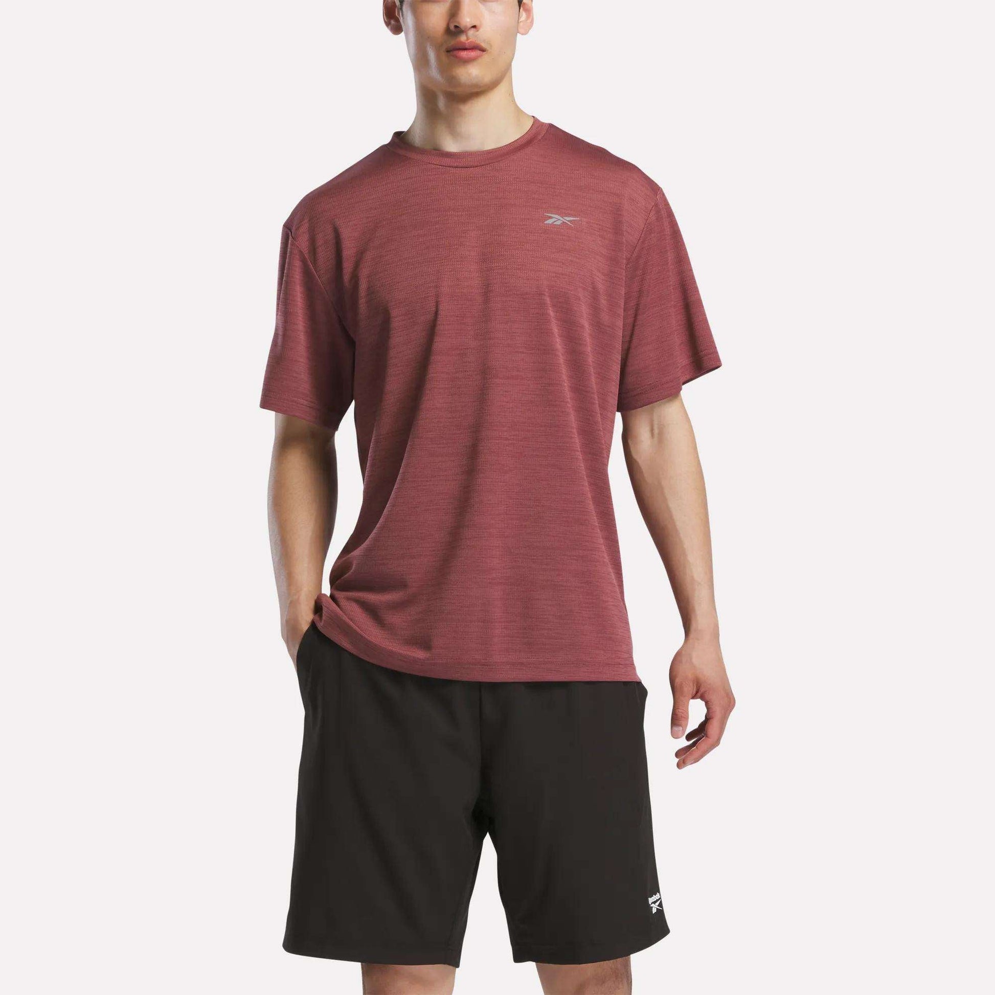 A man wearing a maroon Reebok Men's Director Short Sleeve Top and black shorts stands against a plain white background.