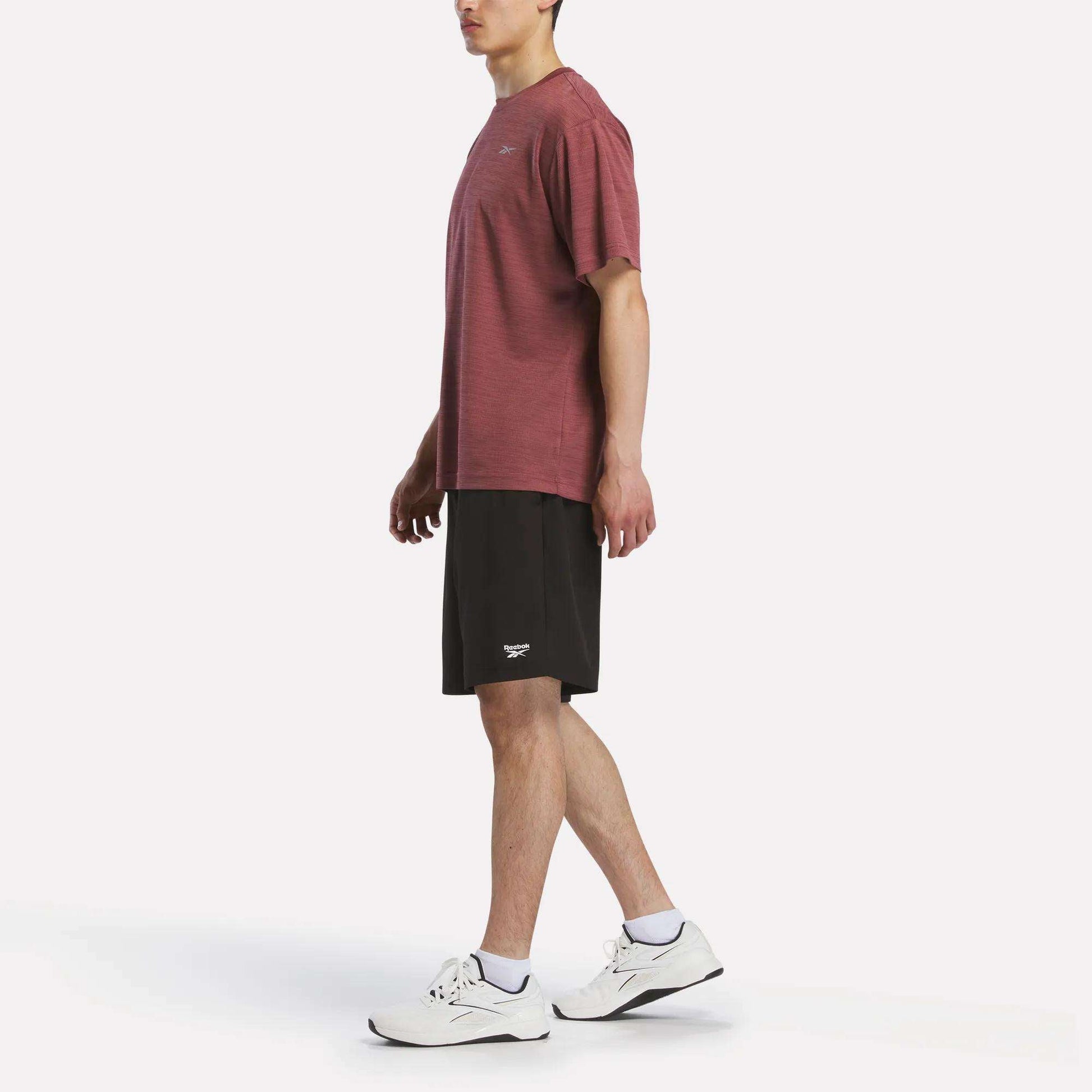 A person stands in profile against a plain white background, wearing a maroon Reebok Men's Director Short Sleeve Top, black shorts, white socks, and white athletic shoes.