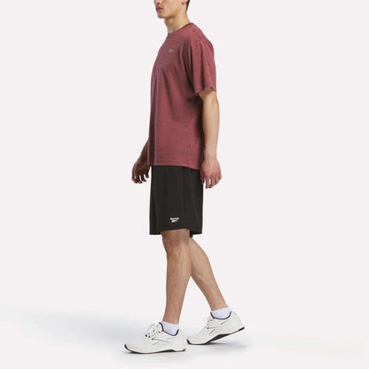 A person stands in profile against a plain white background, wearing a maroon Reebok Men's Director Short Sleeve Top, black shorts, white socks, and white athletic shoes.