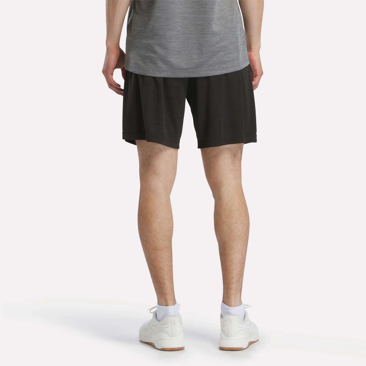 A person is standing with their back to the camera, wearing a gray shirt, white sneakers, and Reebok Men's Inclination Shorts.