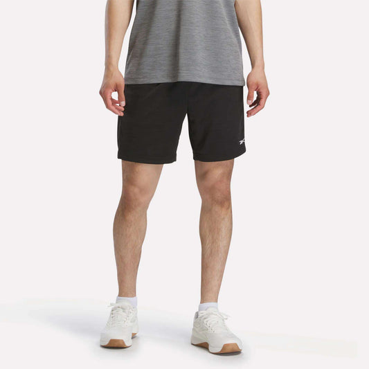 A person wearing a grey t-shirt, black Reebok Men's Inclination Shorts, and white sneakers is standing against a plain white background. The photo shows the person from shoulders to feet.