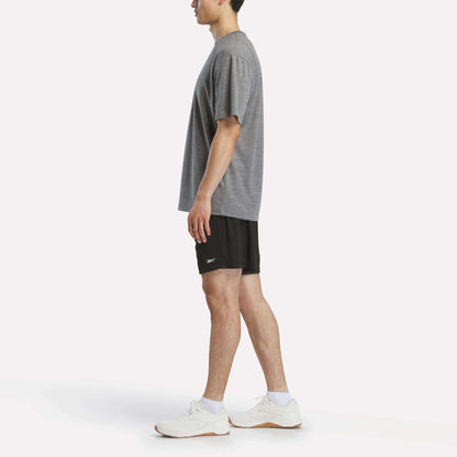 A man stands in profile wearing a loose grey t-shirt, Reebok Men's Inclination Shorts in black, and white athletic shoes against a plain white background.